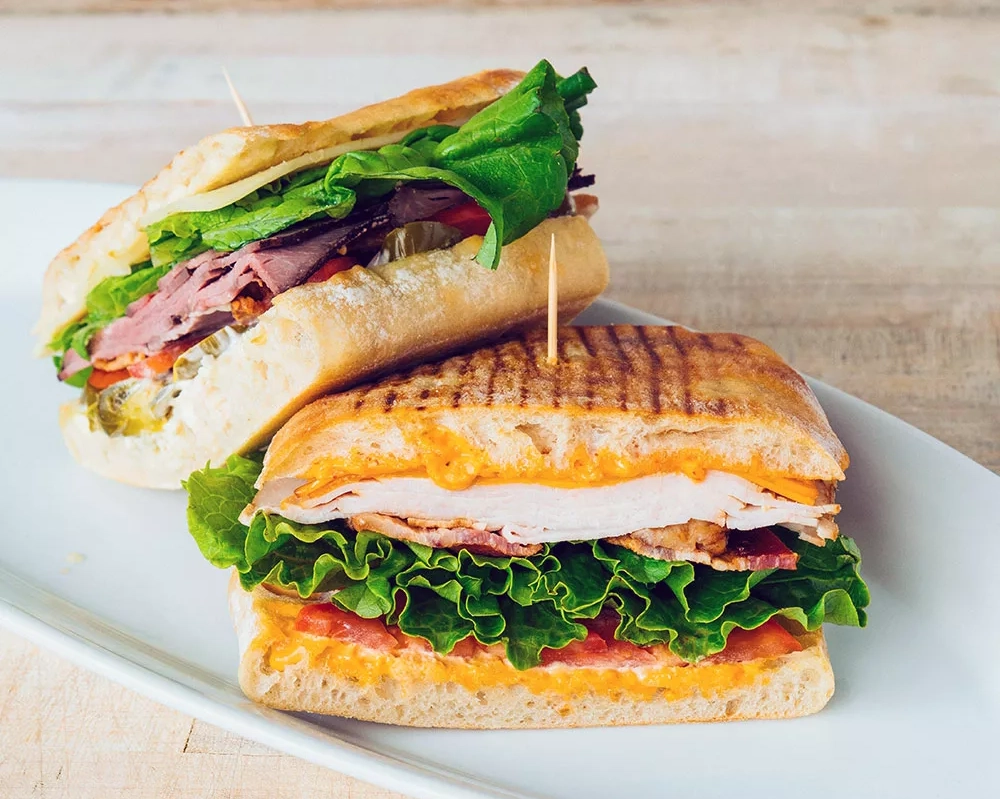 A panini sandwich with cheese, meats, lettuce, and tomatoes on a white plate, showing the concept of easy ready-made meals