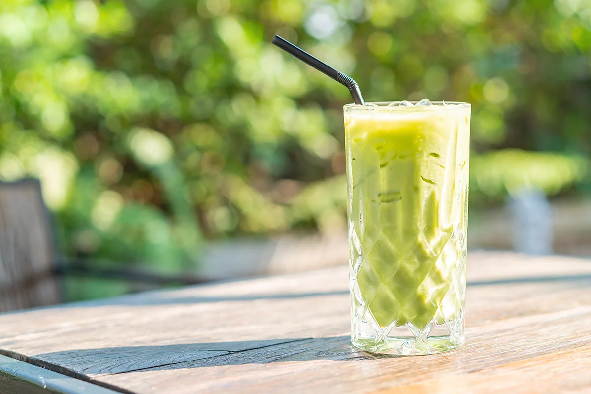A matcha colada cocktail in a glass on a table outside, with leaves in the background