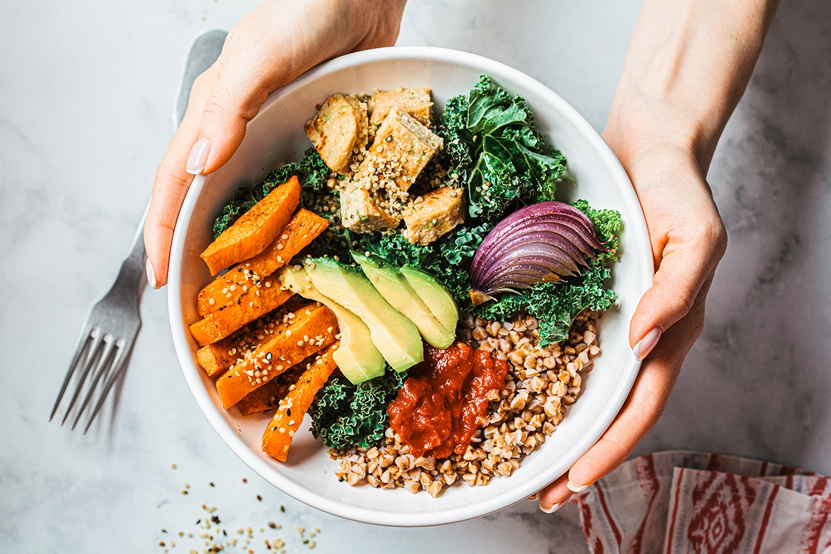 An overhead shot of a salad in a white bowl, with sweet potatoes, kale, avocados, and healthy grains